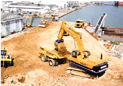 Image of a construction site at a harbour with a large excavator loading dirt into a dump truck, several other construction vehicles nearby, and industrial buildings in the background.