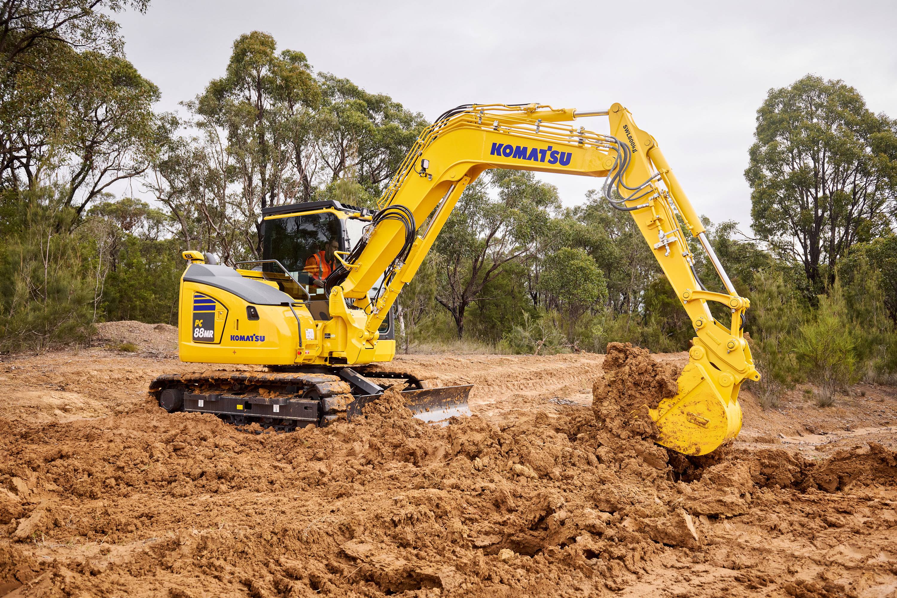 A Komatsu excavator operating in a forested area, digging up soil with its bucket.