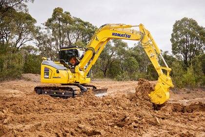 A Komatsu excavator operating in a forested area, digging up soil with its bucket.