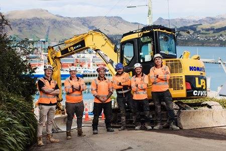 Group of five construction workers in orange safety vests and helmets standing in front of a yellow Komatsu excavator, with a scenic view of a harbor and hills in the background.