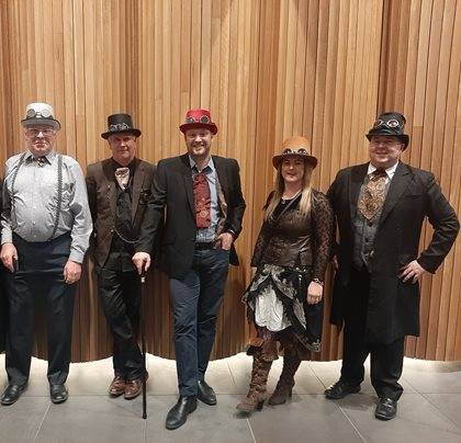 Five people dressed in steampunk attire posing in front of a wooden panel wall.