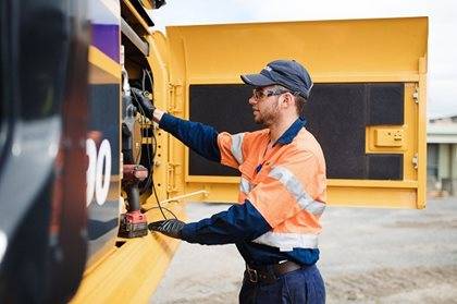 A worker in a high-visibility vest and cap is using a tool on a large yellow heavy machine.