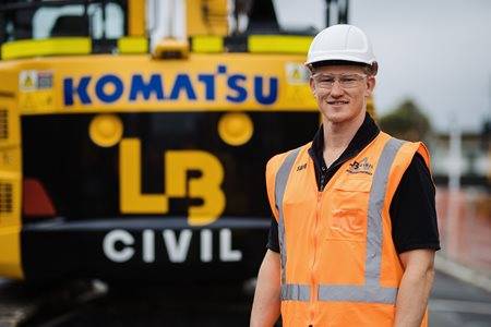 A person in a white hard hat and orange high-visibility vest stands in the foreground, smiling at the camera. Behind them, a large yellow Komatsu excavator with the logo prominently displayed and labeled "LB CIVIL" is visible.