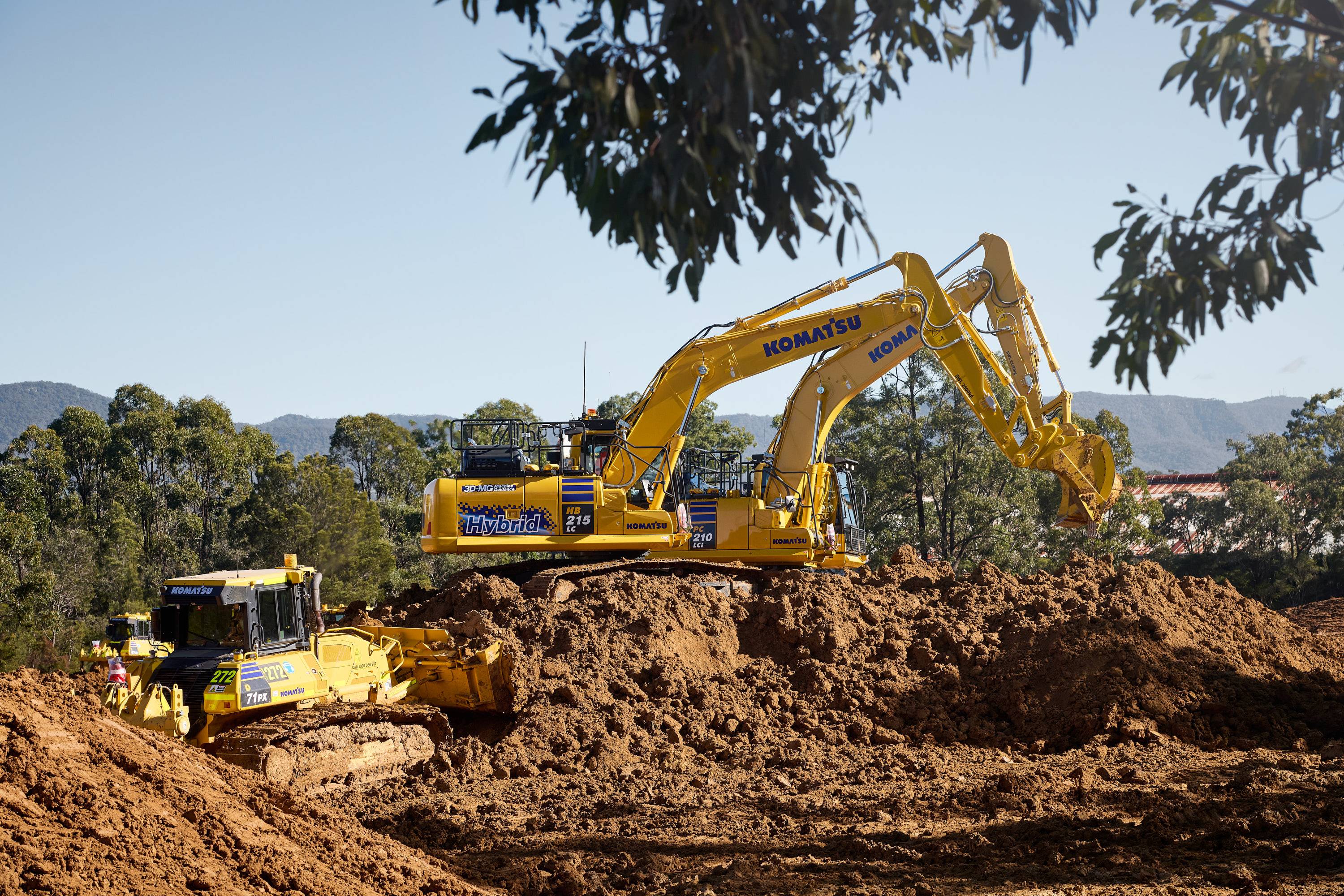 A Komatsu excavator and bulldozer operating on a large dirt mound in a sunny, wooded area.