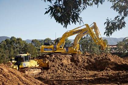 A Komatsu excavator and bulldozer operating on a large dirt mound in a sunny, wooded area.
