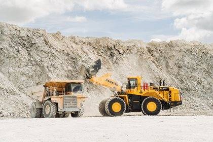 Two large mining trucks are operating in a quarry with a pile of gravel in the background under a cloudy sky.