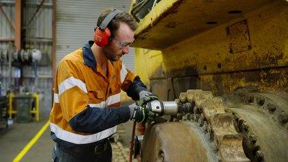 A worker in safety gear repairs heavy machinery in an industrial setting.
